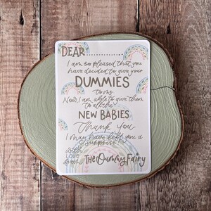 May include: A rectangular card with text, placed on a light green wooden slice. The card features text including "DEAR," "DUMMIES," and "NEW BABIES." Rainbow and floral designs decorate the text. The card is from "The Dummy Fairy."