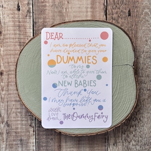 Puede incluir: Una tarjeta blanca con texto y puntos de colores, sobre una rodaja de madera pintada de verde. La tarjeta dice: "DEAR...DUMMIES...NEW BABIES...Thank You...with Love from THE Dummy Fairy."