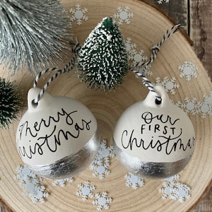 May include: Two white ceramic Christmas baubles with silver accents. One bauble says "Merry Christmas" and the other says "Our First Christmas".