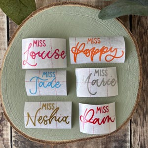 May include: Six rectangular name decals with the word "Miss" followed by a name in various colours. Names include Louise, Poppy, Jade, Marie, Neshia, and Ram. The decals are arranged on a light green wooden slice.