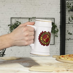 Puede incluir: Jarra de cerveza de cerámica blanca con borde y asa dorados. La jarra presenta un emblema circular con el texto "STONECUTTERS WE DO, WE DO!" en rojo y dorado. El emblema incluye un ancla. La jarra está sostenida por una persona.