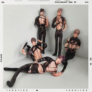 May include: A group of five male figurines in various poses. They are dressed in black leather-like outfits with harnesses and straps. The figurines have different hairstyles and skin tones. The background is a simple gray corner.