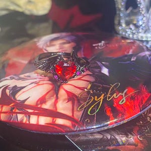 May include: A black ring with a heart-shaped red gemstone and small pink gemstones. The ring has wing-like details on the sides. The ring is on a red and black surface with the word "Sylus" in gold.