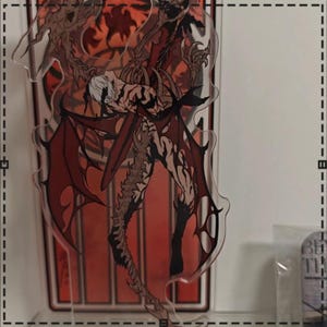 May include: A clear acrylic stand featuring a detailed illustration of a winged demon with red wings and a muscular physique. The artwork has a dark, gothic theme with a red and black color palette.