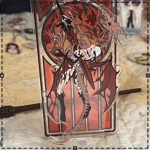 May include: A clear acrylic stand featuring a detailed illustration of a character with red wings and a dragon-like creature. The artwork has a red and orange color scheme with a decorative border. A wand is visible in the background.