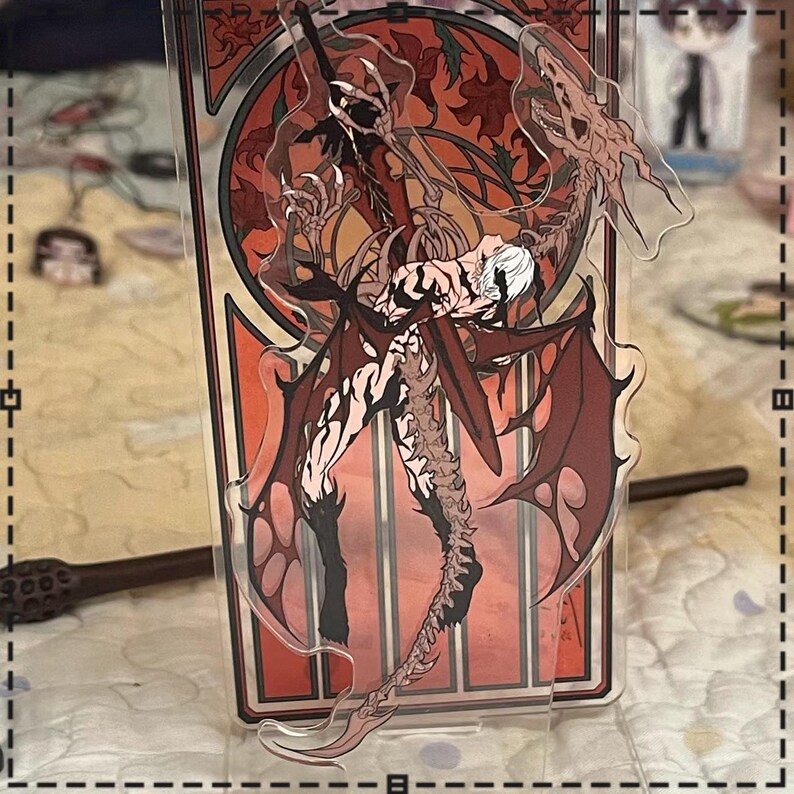 May include: A clear acrylic stand featuring a detailed illustration of a character with red wings and a dragon-like creature. The artwork has a red and orange color scheme with a decorative border. A wand is visible in the background.