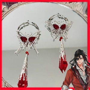 May include: A pair of silver butterfly earrings with red detailing. Each earring has a butterfly design with a red gemstone centre and a dangling, elongated red crystal pendant. The earrings are displayed on a reflective surface.