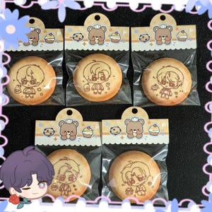 Op de afbeelding: Zes ronde koekjes met cartoonkarakterontwerpen, elk afzonderlijk verpakt. De koekjes zijn lichtbruin met gedetailleerde illustraties. Elk pakket heeft een decoratieve header met een beer, cupcakes en koekjes.