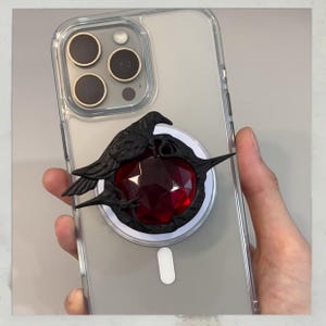 Puede incluir: Una funda transparente para tel&eacute;fono con un dise&ntilde;o de cuervo negro y gema roja. El cuervo est&aacute; posado sobre una gema roja facetada, con un c&iacute;rculo de carga magn&eacute;tica blanco. La funda se sostiene en una mano.