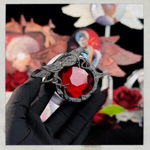 Puede incluir: Un colgante plateado oscuro con una gran gema roja facetada. El colgante est&aacute; adornado con dos figuras de cuervos posadas a ambos lados de la gema. El dise&ntilde;o tiene una est&eacute;tica g&oacute;tica ornamentada, con detalles intrincados y un llamativo contraste de color.