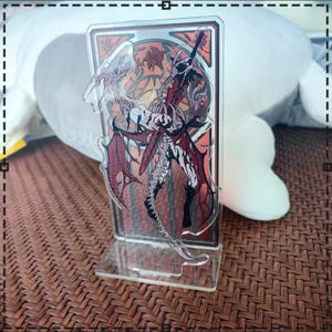 May include: A clear acrylic stand featuring a detailed illustration of a fantasy character with red and black wings, a sword, and a stained-glass style background. The stand is on a clear base.