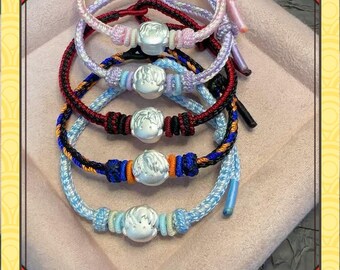 May include: A collection of handmade bracelets in various colors, including light blue, red, and purple. Each bracelet features a silver-colored charm and intricate knotting details. The bracelets are displayed on a soft, pink surface.