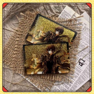 Może przedstawiać: Dwa prostokątne, błyszczące, złote podkładki z żywicy z ilustracją postaci. Postać ma brązowe włosy i nosi czarno-złoty str&oacute;j. Podkładki są umieszczone na powierzchni z juty z wycinkami z gazet i sznurkiem.