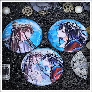 May include: Three circular pin badges with anime-style illustrations. Each badge depicts a different image of two characters with long hair, set against a blue backdrop. The characters are in different poses, with one showing them embracing.