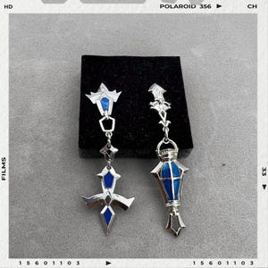 May include: A pair of silver-toned earrings with blue accents. One earring features a cross design, while the other has a lantern shape. Both earrings have intricate details and are displayed on a black surface.