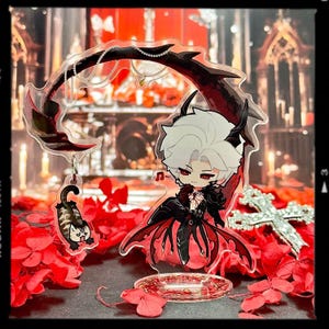 May include: A clear acrylic anime-style figure featuring a character with white hair, red eyes, and a black and red outfit. A small cat charm hangs from the top. The figure is set against a backdrop of red flowers and a cross.