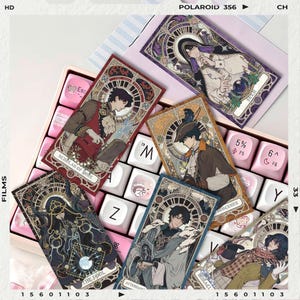 May include: A collection of tarot card designs featuring illustrations of anime-style characters. The cards are in various colors, including red, blue, and gold, and are arranged on a pink and white keyboard. The cards have text such as "Scholar of More" and "The Fool".