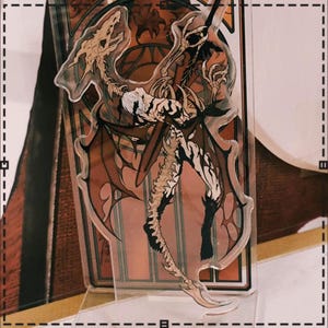May include: A clear acrylic charm featuring a detailed illustration of a skeletal winged creature with a human-like torso. The artwork incorporates shades of brown, black, and white, with a gothic aesthetic. The charm is designed with intricate details.