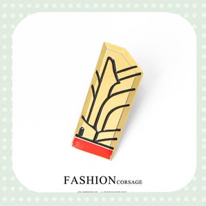 May include: A gold-colored rectangular brooch with black line art and a red accent at the bottom. The brooch has a geometric design and the text "FASHION CORSAGE" is below it.