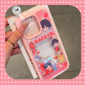 May include: A pink phone case with a clear central section and cartoon illustrations. The case has the name "RAFAYEL" and various cartoon characters, including a fish, a dolphin, and a dragon. The case is held by a hand.