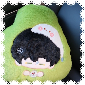 May include: A plush avocado-shaped pillow with a cartoon character design. The character has black hair, a white hat with pink snowflake accents, and is wearing a green outfit. A small snowman is embroidered at the bottom.