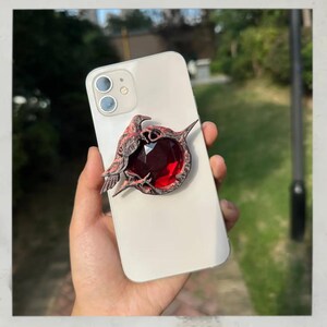 Puede incluir: Un soporte decorativo para tel&eacute;fono con una joya roja oscura rodeada por un dise&ntilde;o detallado de p&aacute;jaro y espinas de color marr&oacute;n rojizo. El soporte est&aacute; adherido a un tel&eacute;fono inteligente blanco, ofreciendo una est&eacute;tica &uacute;nica y un mejor manejo.