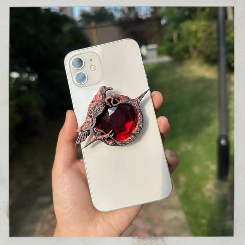 Puede incluir: Un soporte decorativo para tel&eacute;fono con una joya roja oscura rodeada por un dise&ntilde;o detallado de p&aacute;jaro y espinas de color marr&oacute;n rojizo. El soporte est&aacute; adherido a un tel&eacute;fono inteligente blanco, ofreciendo una est&eacute;tica &uacute;nica y un mejor manejo.