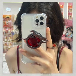Puede incluir: Una funda transparente para tel&eacute;fono con un agarre para tel&eacute;fono de metal oscuro con forma de cuervo y gema roja. El cuervo est&aacute; posado en un marco circular con una gran gema roja en el centro.