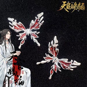 Può includere: Due spille a forma di farfalla con montatura argentata e ali in smalto rosso. Sono esposte su una superficie nera. L'immagine include anche un disegno di una persona in abiti bianchi e caratteri cinesi.