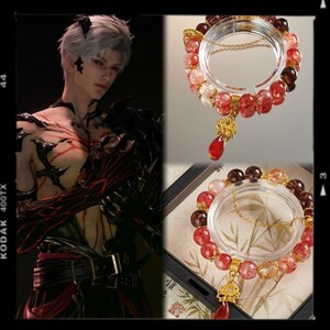 May include: A bracelet with red and clear beads, a gold-coloured charm, and a red teardrop pendant. The bracelet is displayed on a clear circular stand. The image also features a person with white hair and black markings.