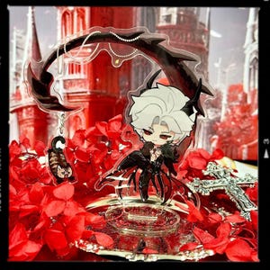 May include: A clear acrylic charm featuring a chibi anime character with white hair, red eyes, and a black outfit. The charm has a decorative black element and a small cat charm. The background is a red floral arrangement.