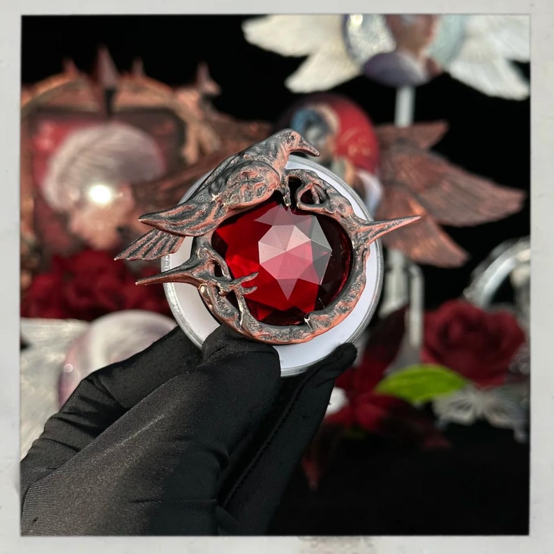 Puede incluir: Un espejo compacto decorativo de estilo antiguo con una gema roja facetada en forma de coraz&oacute;n. El marco de metal color bronce est&aacute; adornado con un dise&ntilde;o de p&aacute;jaro y rama. El espejo compacto es sostenido por una mano enguantada sobre un fondo oscuro.