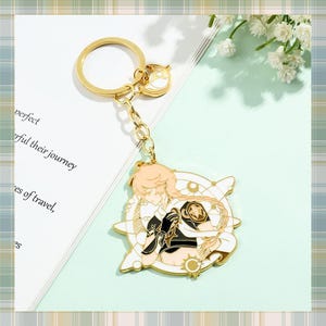 Puede incluir: Un llavero dorado con un diseño de personaje en blanco, negro y beige. El llavero tiene un anillo circular y una cadena, con un dije decorativo. El fondo incluye texto y flores.
