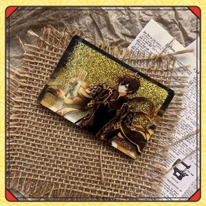 Może przedstawiać: Prostokątny akrylowy urok z ilustracją postaci i złotym brokatem. Postać jest ubrana w ciemne ubrania i ma brązowe włosy. Urok jest umieszczony na tle juty i gazety. Widoczny jest napis 'jrboa'.