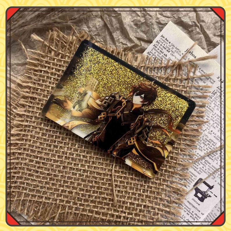 Może przedstawiać: Prostokątny akrylowy urok z ilustracją postaci i złotym brokatem. Postać jest ubrana w ciemne ubrania i ma brązowe włosy. Urok jest umieszczony na tle juty i gazety. Widoczny jest napis 'jrboa'.