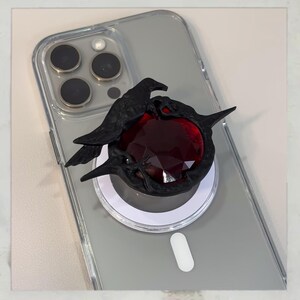 Puede incluir: Soporte de tel&eacute;fono negro con un dise&ntilde;o de cuervo y una gema roja. El cuervo est&aacute; detallado, con alas y picos. El soporte est&aacute; adherido a una funda transparente para tel&eacute;fono.