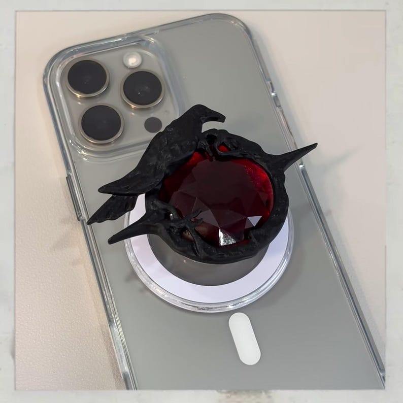 Puede incluir: Soporte de tel&eacute;fono negro con un dise&ntilde;o de cuervo y una gema roja. El cuervo est&aacute; detallado, con alas y picos. El soporte est&aacute; adherido a una funda transparente para tel&eacute;fono.