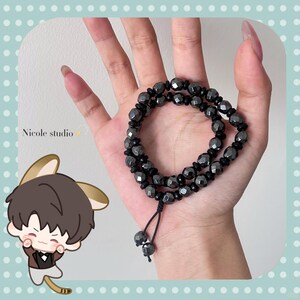 May include: A dark gray beaded bracelet with a black cord and two dangling beads. The bracelet is held in a hand. The image also includes a cartoon character and the text "Nicole studio".