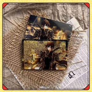 Może przedstawiać: Dwa prostokątne elementy artystyczne z żywicy przedstawiające ilustrację postaci z tłem ze złotego brokatu. Postać jest przedstawiona w ciemnym stroju z brązowymi włosami. Elementy są umieszczone na tle z juty i papieru.