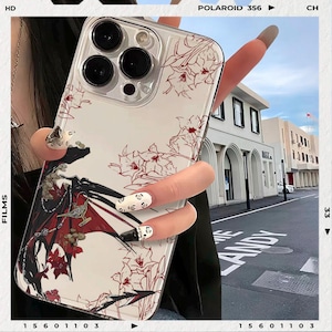 May include: Clear phone case with a white background and a red floral design. The case features a decorative illustration with a figure and a red and black color scheme. The case is held in a hand.