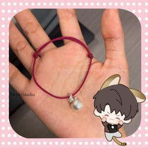 May include: A burgundy-colored bracelet with a pearl charm. The bracelet is made of a woven cord and features a small, round pearl pendant. The bracelet is displayed on a person's hand. The text "Nicole studio" is visible.