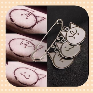 May include: A silver-toned safety pin with three cat-shaped charms. The charms are light gray with white cat faces. The image also shows three drawings of cat faces on a light brown surface.