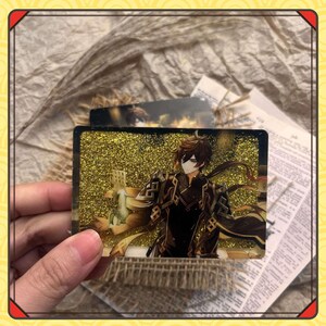 Może przedstawiać: Prostokątna karta przedstawiająca postać z anime z brązowymi włosami i czarnym strojem. Tło to złoty brokat. Karta jest trzymana w dłoni, z innymi kartami i papierem w tle.