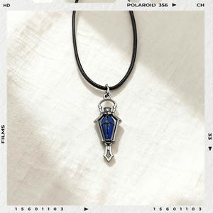Könnte beinhalten: Halskette mit silberfarbenem Anhänger und Sarg-förmigem Anhänger in Blau und Silber. Der Anhänger hängt an einem schwarzen Band. Der Hintergrund ist eine weiße, strukturierte Oberfläche.