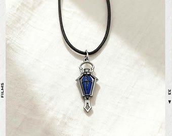 May include: A silver-toned pendant necklace with a blue and silver coffin-shaped charm. The charm has a blue center and is suspended from a black cord. The background is a white, textured surface.