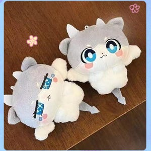 May include: Two soft toy figures resembling cartoon animals. They have grey heads, white bodies, and large blue eyes with pink cheeks. The toys have small arms and a tail. One is lying down, and the other is standing.