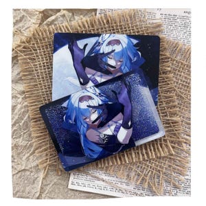 May include: Two rectangular cards featuring a blue-haired anime character in a dark outfit, with a glittery overlay. The character is depicted with closed eyes and gloved hands. The cards are placed on a textured, neutral-toned surface.