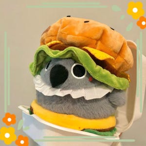 May include: A plush koala wearing a burger costume. The koala is gray with black eyes and a white ruffled collar. The burger has a yellow bun, green lettuce, and a brown patty. The koala is in a white container.