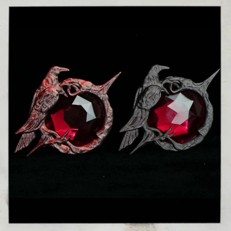 Puede incluir: Dos broches decorativos con un dise&ntilde;o de cuervo que sostiene una gema roja facetada. El cuervo y el engaste tienen un acabado met&aacute;lico. Los broches se muestran sobre un fondo negro.