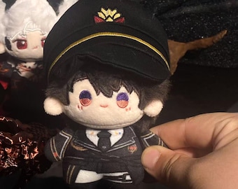 May include: A plush doll wearing a black uniform and hat with gold trim and a decorative emblem. The doll has black hair, and a white face with red and purple accents. Another doll is visible in the background.